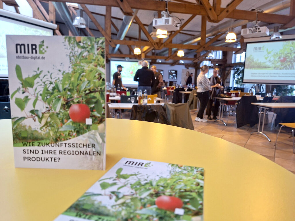 Veranstaltungsraum mit Broschüre über regionale Produkte auf gelbem Tisch, im Hintergrund Menschen und Präsentationsbildschirm.
