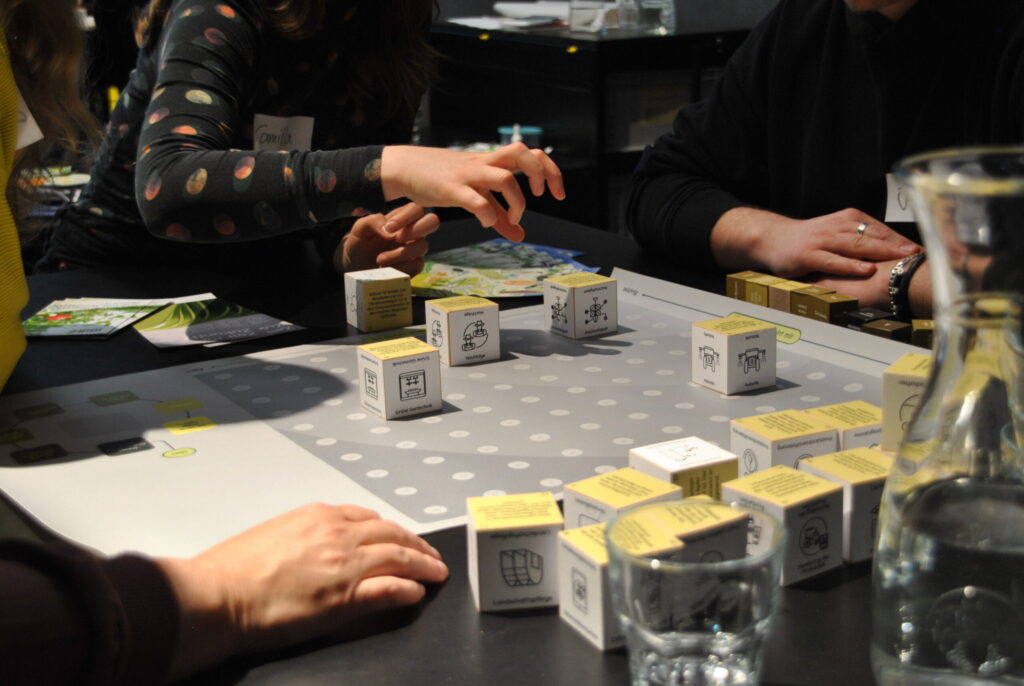 Personen spielen ein Brettspiel mit bedruckten Würfeln auf einem Tisch, im Hintergrund liegen Handzettel.